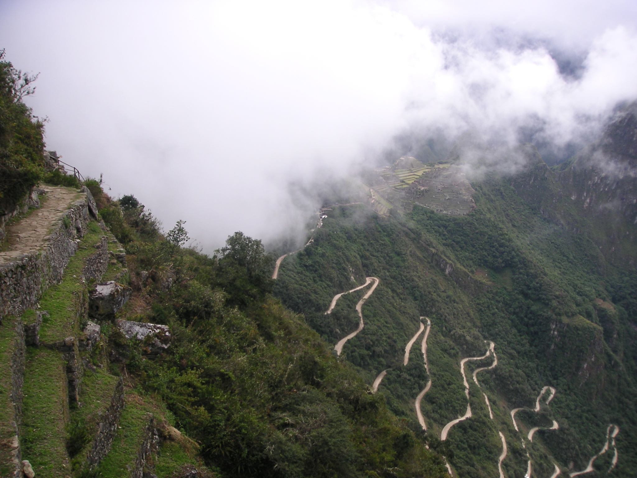 Machu Picchu is covered in clouds. The winding dirt and gravel bus road is seen below Machu Picchu.